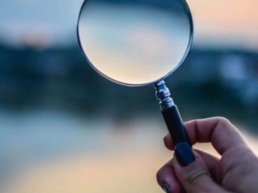 A hand holding a magnifying glass against a soft, blurred sunset background, bringing the center of the scene into subtle focus.
